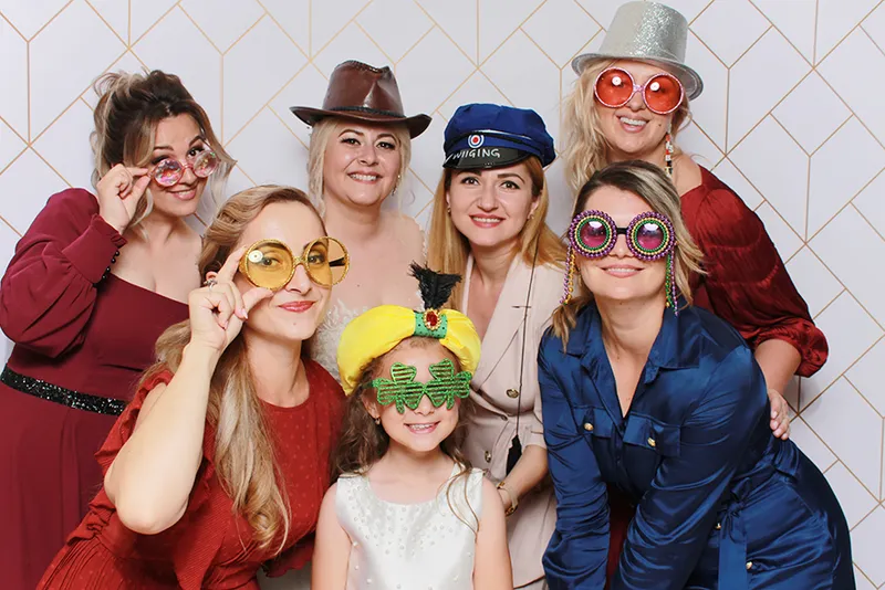 Grup de femei și o fetiță cu ochelari și pălării haioase la cabina foto, la o nuntă